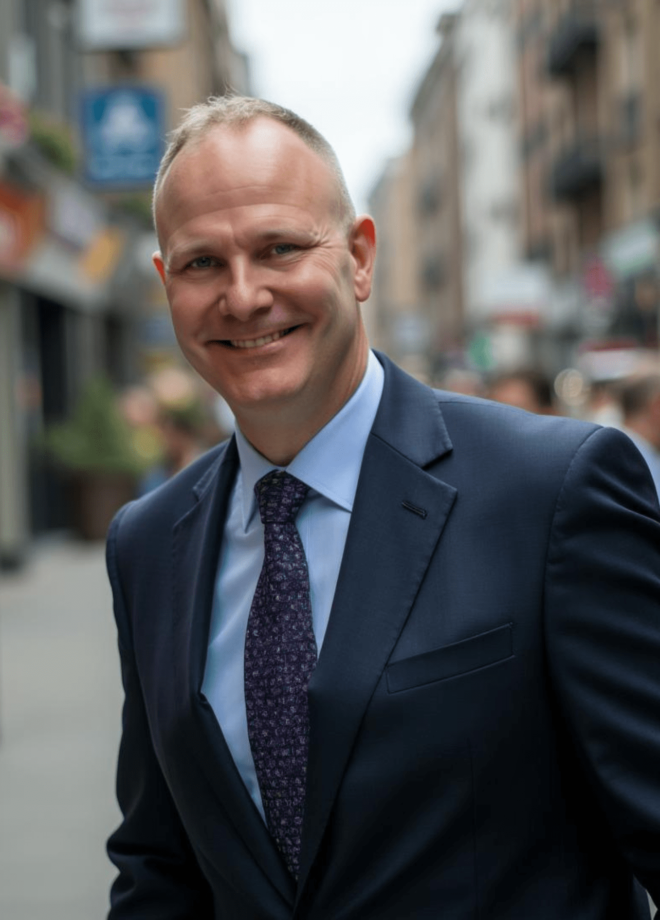 A smiling bald man sits with his hands clasped on a dark surface, wearing a light green collared shirt, against a black background.