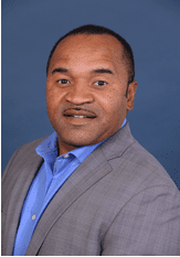 A man in a gray suit jacket and blue shirt poses in front of a solid blue background.