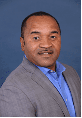 A man in a gray suit jacket and blue shirt poses in front of a solid blue background.